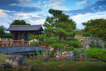 Himeji, Japan - Sep 24 2024, panoramic view of Sakuramon Bridge and entrance gate to Himeji Castle, White Heron Castle, with a tree in foreground, at daytime with cloudy sky, Himeji, Japan