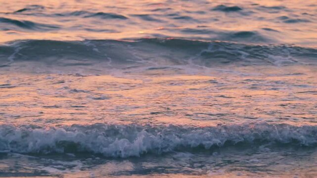 Energetic close-up of foamy ocean waves crashing onto the beach at sunset. Shot on tripod, this scene captures the glowing reflections of pink and golden light on the moving water.