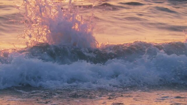 Close-up tripod shot of a vivid ocean wave splashing onto the shore at sunset. The warm golden-pink light reflects beautifully on the water, highlighting the motion and texture of the sea.