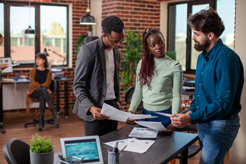 Black female entrepreneur with papers in hand, explains business project plans to male employees in startup office. Businesswoman holds and gestures toward documents during discussion with coworkers.