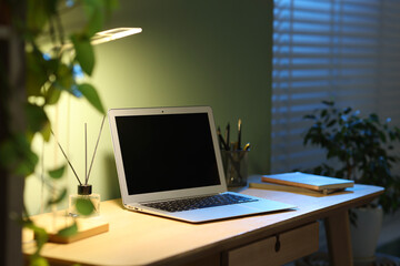 Fototapeta premium Comfortable workplace with laptop and lamp on wooden desk at home office in evening
