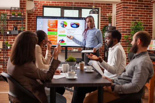 Black female analyst points to screen displaying business analytics, giving explanations to diverse employees seated at table. Businesswoman with clipboard, leading team meeting in brick wall office.
