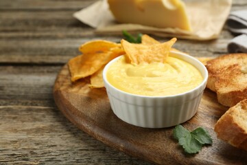 Tasty cheese dipping sauce in bowl, nacho chips and croutons on wooden table, closeup