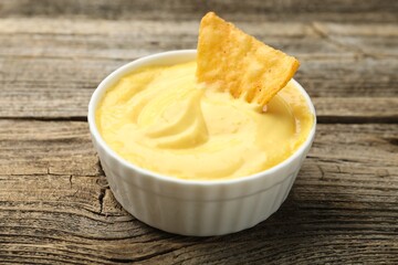 Tasty dipping cheese sauce in bowl and nacho chip on wooden table, closeup