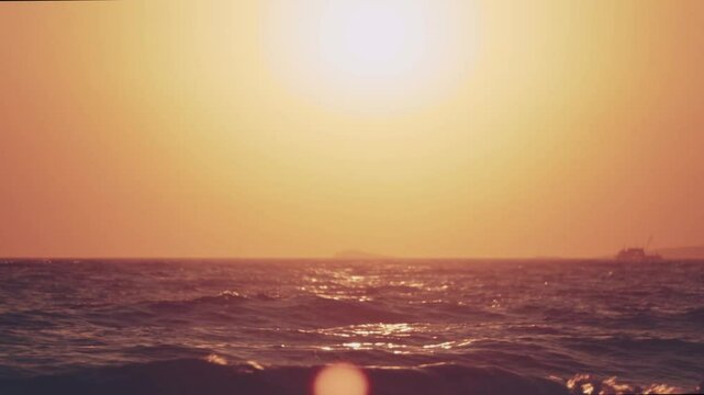 Minimalist tripod shot of the glowing sun above a calm ocean horizon. Gentle waves shimmer with golden reflections in the warm evening light, while a distant ship silhouette adds depth to composition