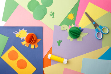 DIY 3D paper dinosaur crafts with colorful craft supplies on a multicolored background. Flat lay...
