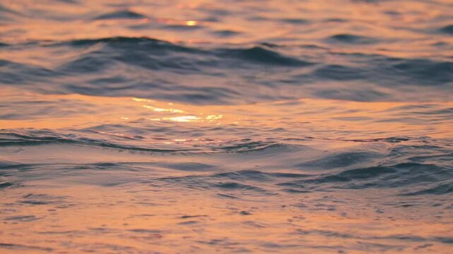 Detailed tripod shot of a small wave curling and breaking gently along the shore during golden hour. The warm sunset light reflects on the water&rsquo;s surface, creating soft pink and gold hues.