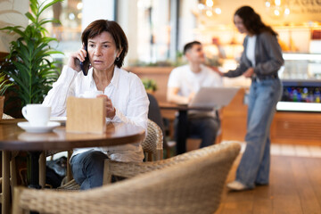 Mature woman talking on mobile phone while sitting at table in coffee shop. European woman answers a business call in a cafe and drinks hot coffee