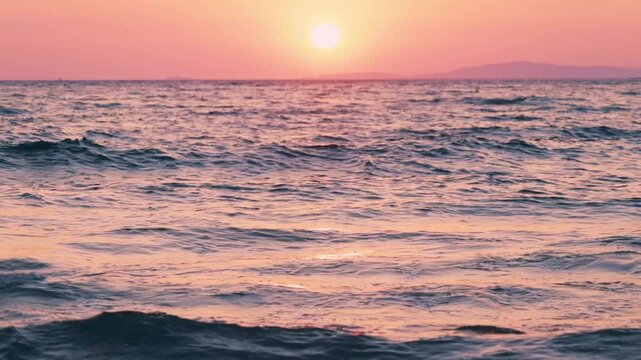 Tripod shot of soft waves washing onto the beach under a delicate pastel sunset sky. The sun nears the horizon, casting pink and lavender tones across the sea, evoking a romantic coastal atmosphere