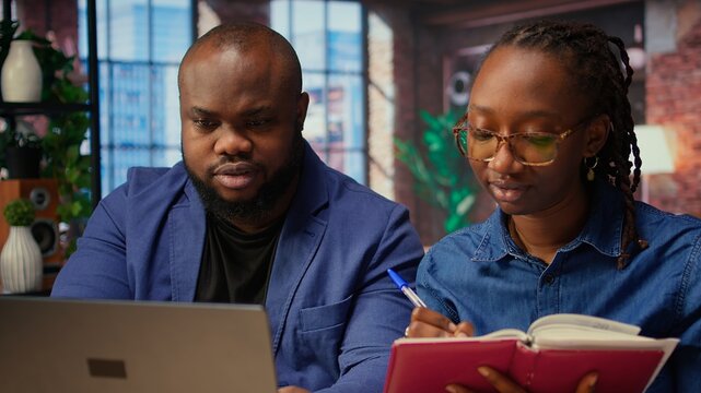 Black remote employees organizing calendar appointments using laptop and ledger, checking papers and synchronizing important tasks on schedule. Man and woman review files at home. Camera A.