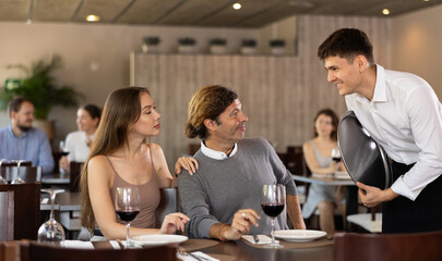 Young guy waiter taking order from couple adult man and young woman in restaurant