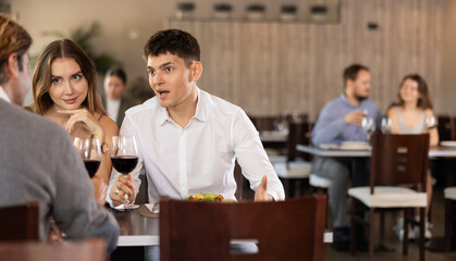 Couple young man and woman with adult man eat drink wine and chat in restaurant