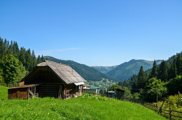 Scenic view of a wooden countryside cabin amidst a lush green meadow, surrounded by forested mountains under a clear blue sky. Carpathians, Ukraine