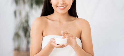 Happy asian woman wrapped in towel using face or body nourishing cream, bedroom interior. Pretty young lady using moisturizer for her face or body after shower in the morning, copy space