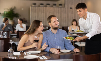 Young guy waiter brings order from couple young man and young woman in restaurant