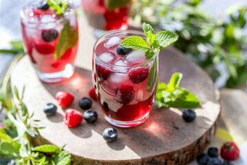 Vibrant glasses filled with iced berry beverages display a mix of fresh raspberries and blueberries. Lush foliage surrounds these refreshing drinks, capturing the essence of summer.