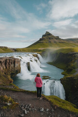 A solitary traveler in a pink jacket gazes in awe at the cascading waterfall in a remote canyon. The serene waters flow as green hills rise majestically, creating a stunning backdrop.