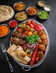 A beautifully arranged platter of grilled meat skewers, roasted vegetables, flatbread, and fresh herbs is served alongside colorful salads and various dipping sauces on a wooden table.