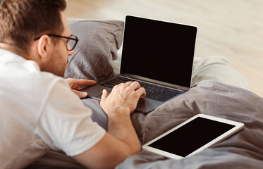 Man With Laptop And Digital Tablet Working From Home Lying On Stomach In Bed Indoors. Freelance Lifestyle Concept. Mockup, Back-View