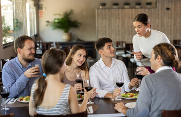 Young woman waitress taking order from group of friends at table in restaurant