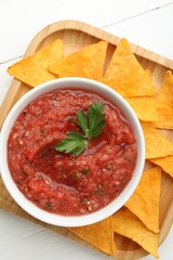 Delicious spicy salsa sauce in bowl and nacho chips on white wooden table, top view