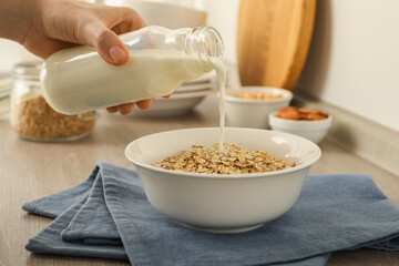 Woman pouring milk into bowl with oatmeal at wooden table, closeup