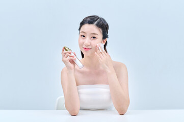 A young Asian female model wearing a tube top is sitting in front of a white background, applying...