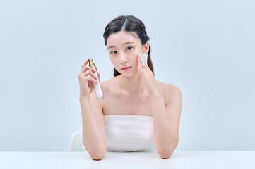 A young Asian female model wearing a tube top is sitting in front of a white background, applying...
