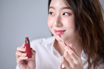 A woman with long hair, wearing a white shirt, is happily applying lip gloss while doing her makeup