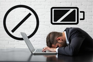 Exhausted businessman sleeping on laptop with low battery icons on white brick wall.