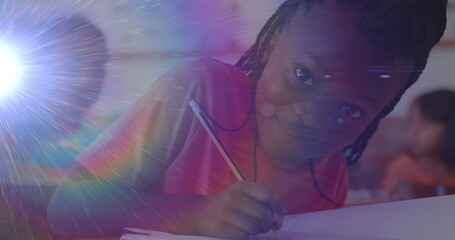 Image of portrait of happy biracial schoolgirl writing during lesson at school smiling to camera