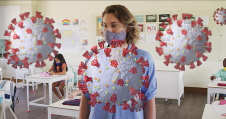 Image of covid 19 cells and female teacher and children in classroom wearing face masks