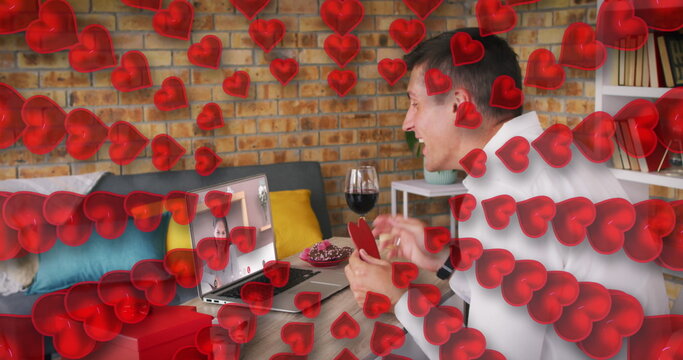 Heart icons against caucasian man holding a heart shaped placard while having a image call on laptop