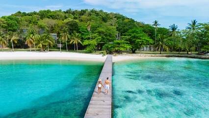 A couple enjoys a serene walk along a wooden pier on Koh Kham Island in Thailand. The turquoise waters and palm trees create a blissful tropical atmosphere perfect for relaxation.
