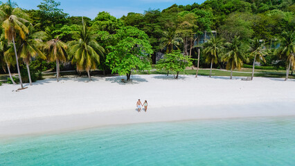 Two people stroll along the pristine white sands of Koh Kham Thailand, framed by swaying palm trees...