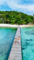 A couple strolls hand in hand on a wooden pier, surrounded by clear turquoise waters and lush green palm trees. at Koh Kham Island Thailand, drone view