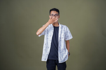 Young Asian man with casual glasses showing surprised expression, covering his mouth with his hand....