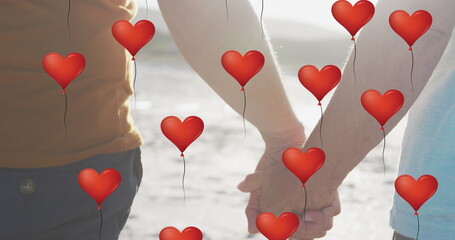 Image of red heart love balloons digital icons over couple holding hands on beach