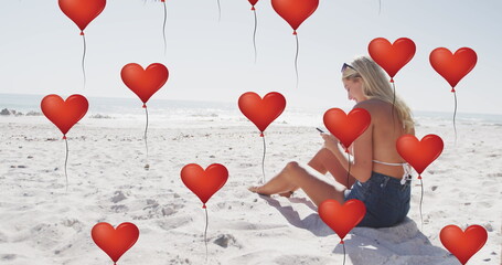 Image of red heart love balloons digital icons over woman using smartphone on beach