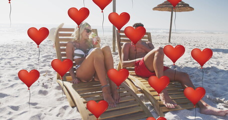 Image of red heart love balloons digital icons over couple in deckchairs with drinks on beach