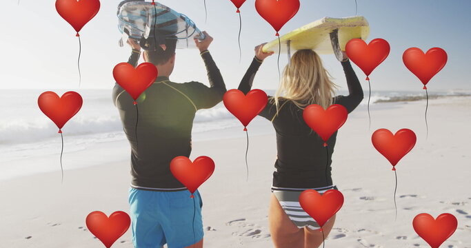 Image of red heart love balloons digital icons over couple carrying surfboards on beach