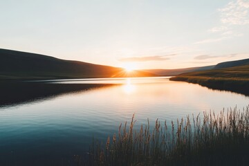 Obraz premium A tranquil lake captures the orange and pink hues of sunset, surrounded by green hills and gently swaying reeds in the foreground, creating a peaceful atmosphere