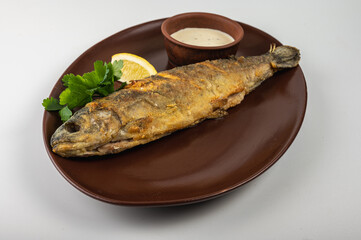 Crispy fried whole fish served on a brown plate with fresh parsley, lemon wedge and creamy sauce. A delicious seafood dish, perfect for a restaurant menu or food blog