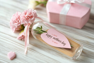 A heartfelt Mother's Day gift: delicate pink carnations nestled in a glass bottle, a sweet message of love, accompanied by a pink gift box on a soft white wooden surface.