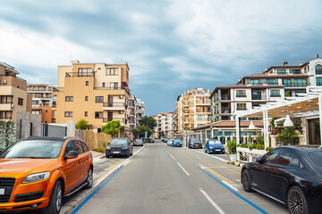 Fototapeta premium Urban street scene with parked cars and residential buildings under cloudy sky. Bulgaria, Pomorie