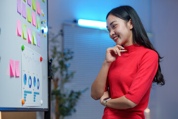 Young professional in a red top analyzing charts and sticky notes on a whiteboard, showcasing a business strategy meeting in a modern office environment