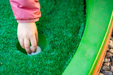 Child Hand Retrieving Golf Ball from Hole on Mini-Golf Course. Little girl playing mini golf outdoor. Family vacations or resort.