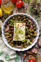 Feta cheese with mixed olives, olive oil and aromatic herbs in a casserole dish on a wooden table, top view.