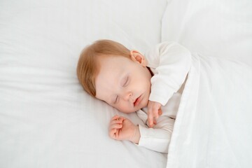 Baby boy sleeping under a blanket in a white bodysuit on the bed with his hand folded under his cheek, sweet healthy baby sleep at home