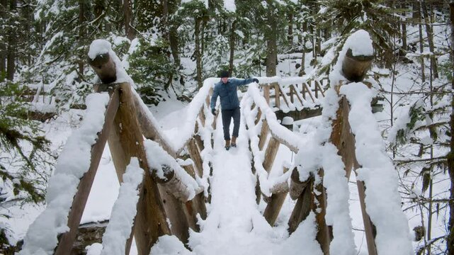 Man Exploring Snowy Forest Crossing Wood Footbridge